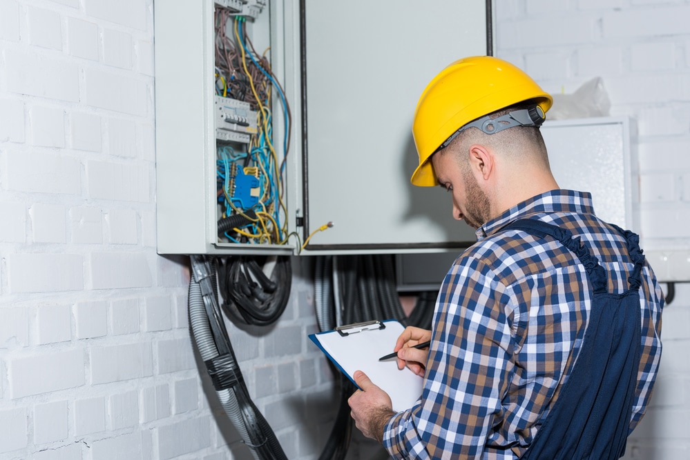 A man in a hard hat is holding a clipboard in front of an electrical box.