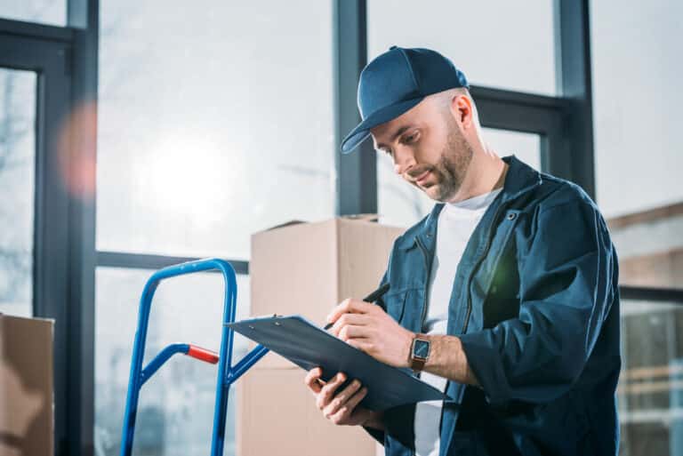 A man is writing on a clipboard in front of boxes.