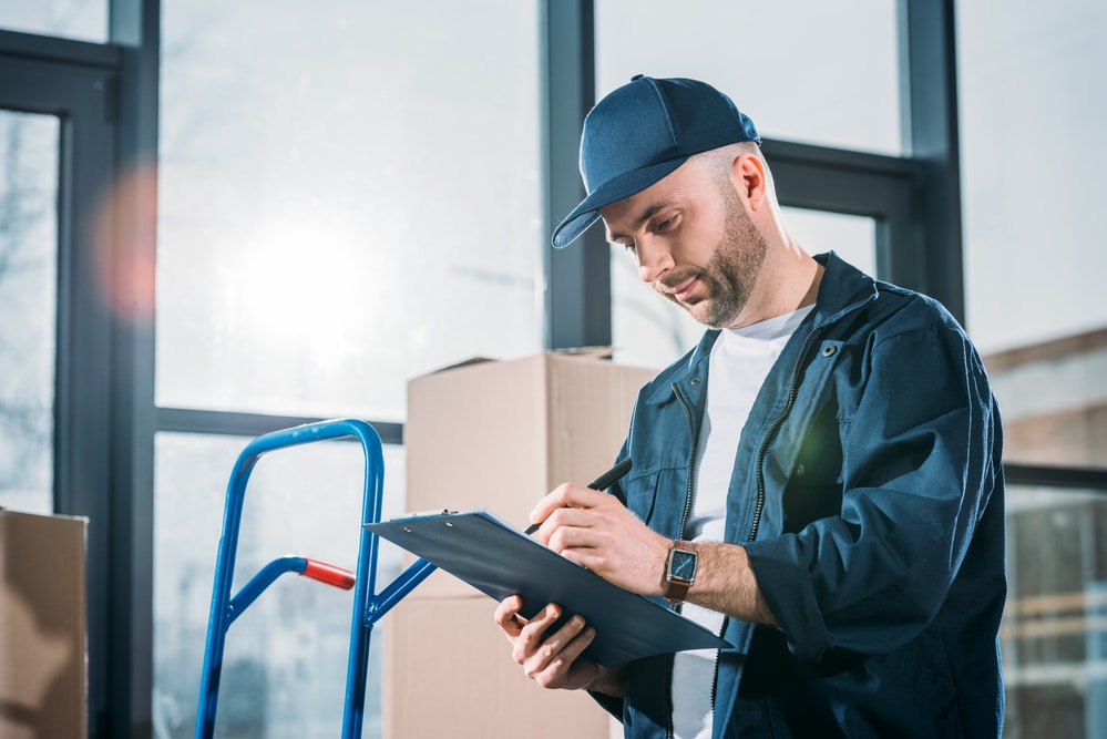 A man is writing on a clipboard in front of boxes.