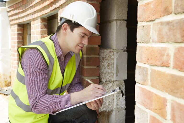 A construction worker writing on a clipboard in front of a brick wall.
