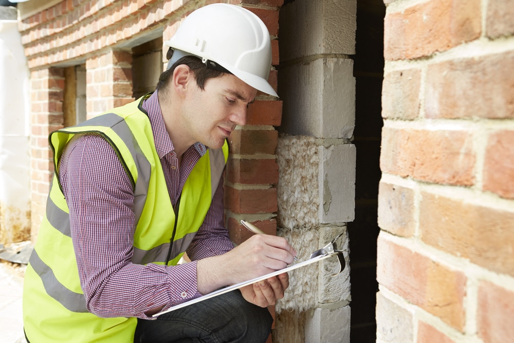 A construction worker writing on a clipboard in front of a brick wall.