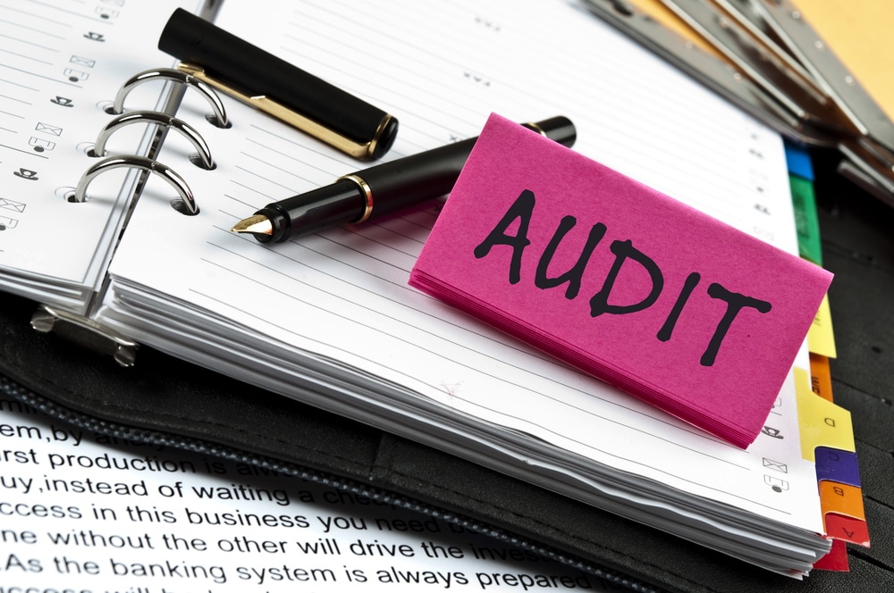 A pink sticky note with the word audit on it sits on top of a notebook filled with medical documents and records