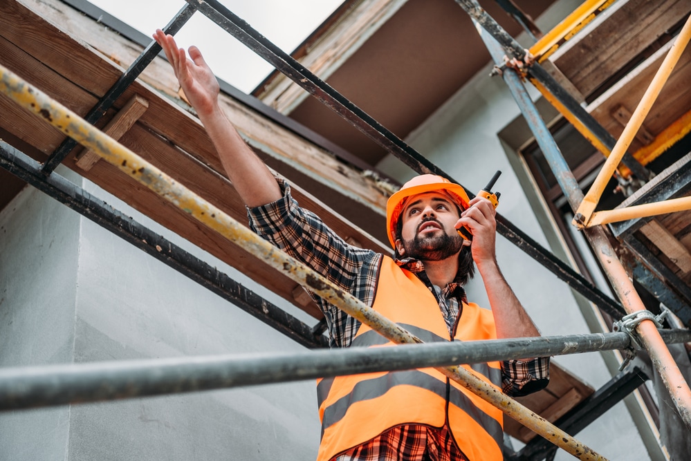 A construction worker talking on a walkie talkie while standing on scaffolding.
